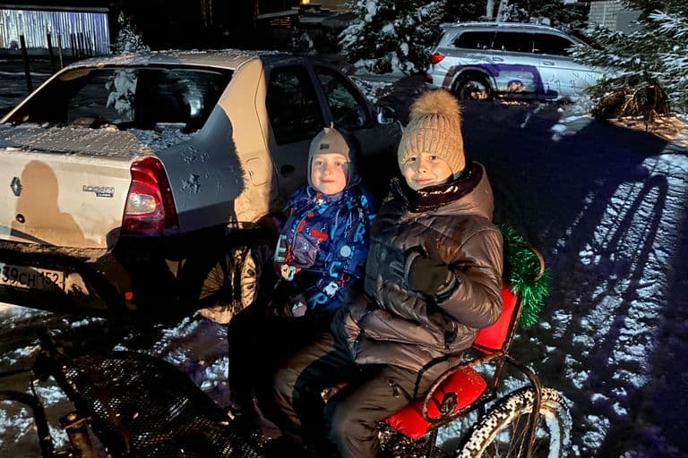 Двое детей в зимней одежде сидят в санях у машины, зимний вечер