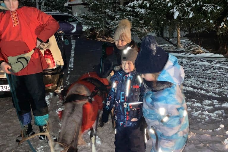 Дети и взрослый рядом с северным оленем, запряжённым в сани, зимний вечер